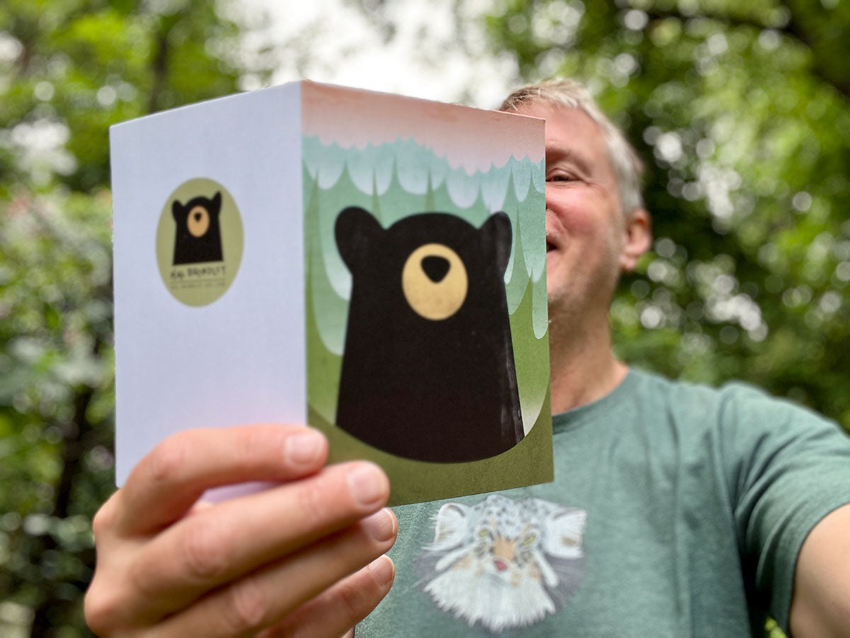 A man outdoors holds up a small greeting card featuring a stylized black bear face and a green forest background. The back of the card has a circular black bear logo and the signature 'Hal Brindley' below it.